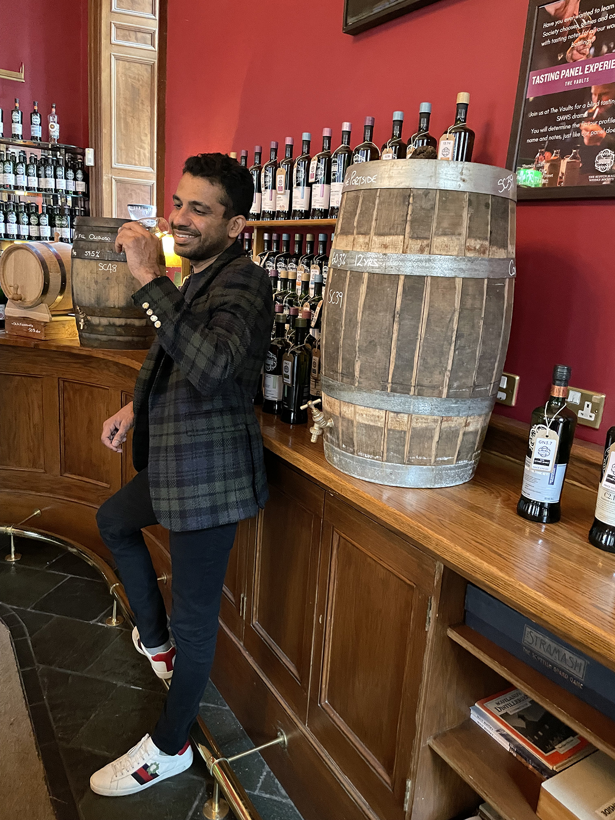 A man in a tartan jacket drinking whiskey next to a cask in a whiskey shop