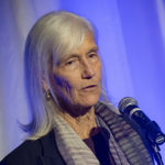 A woman with grey hair speaking into a microphone with a purple backdrop