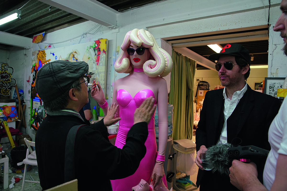 A man in a hat and jacket saying hello to a plastic blonde woman in a pink dress