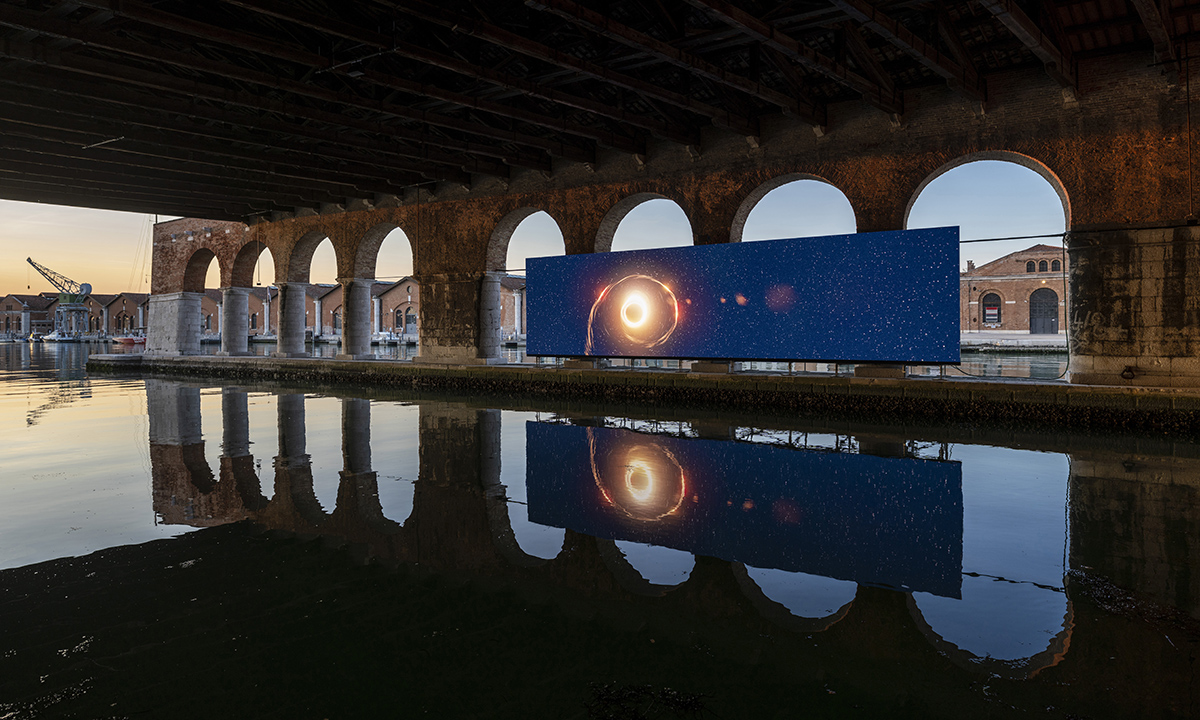 A screen on the river underneath arches and the reflection in the water of a galaxy