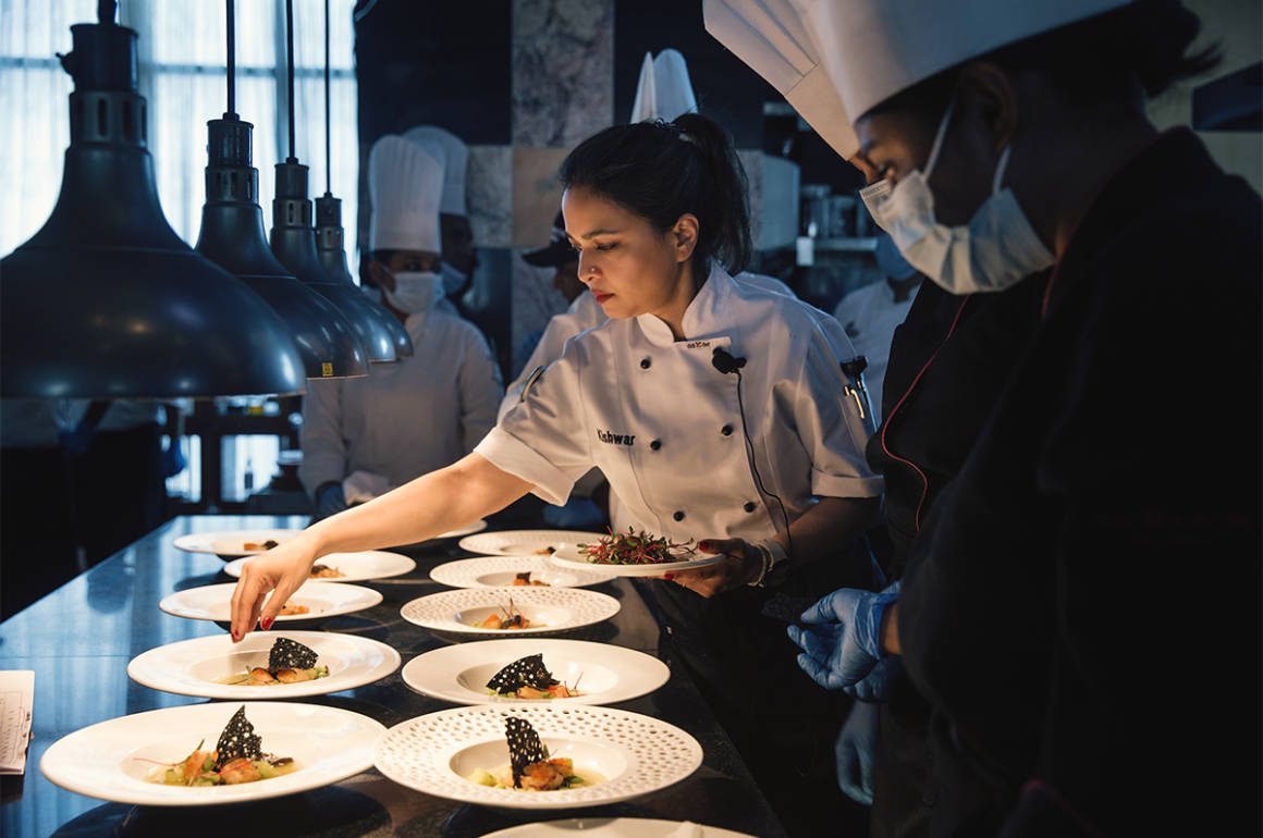 Kishwar Chowdhury showing a chef wearing a top hat how to prepare many plates of food
