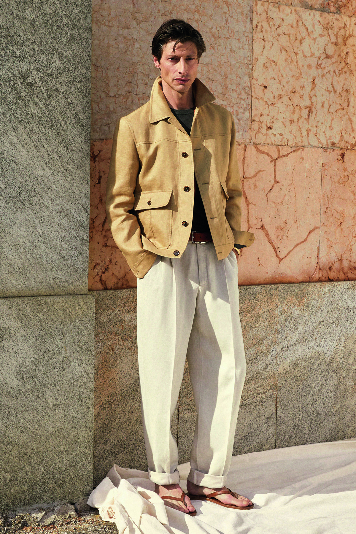A man wearing white trousers and a cream jacket, standing by a stone wall