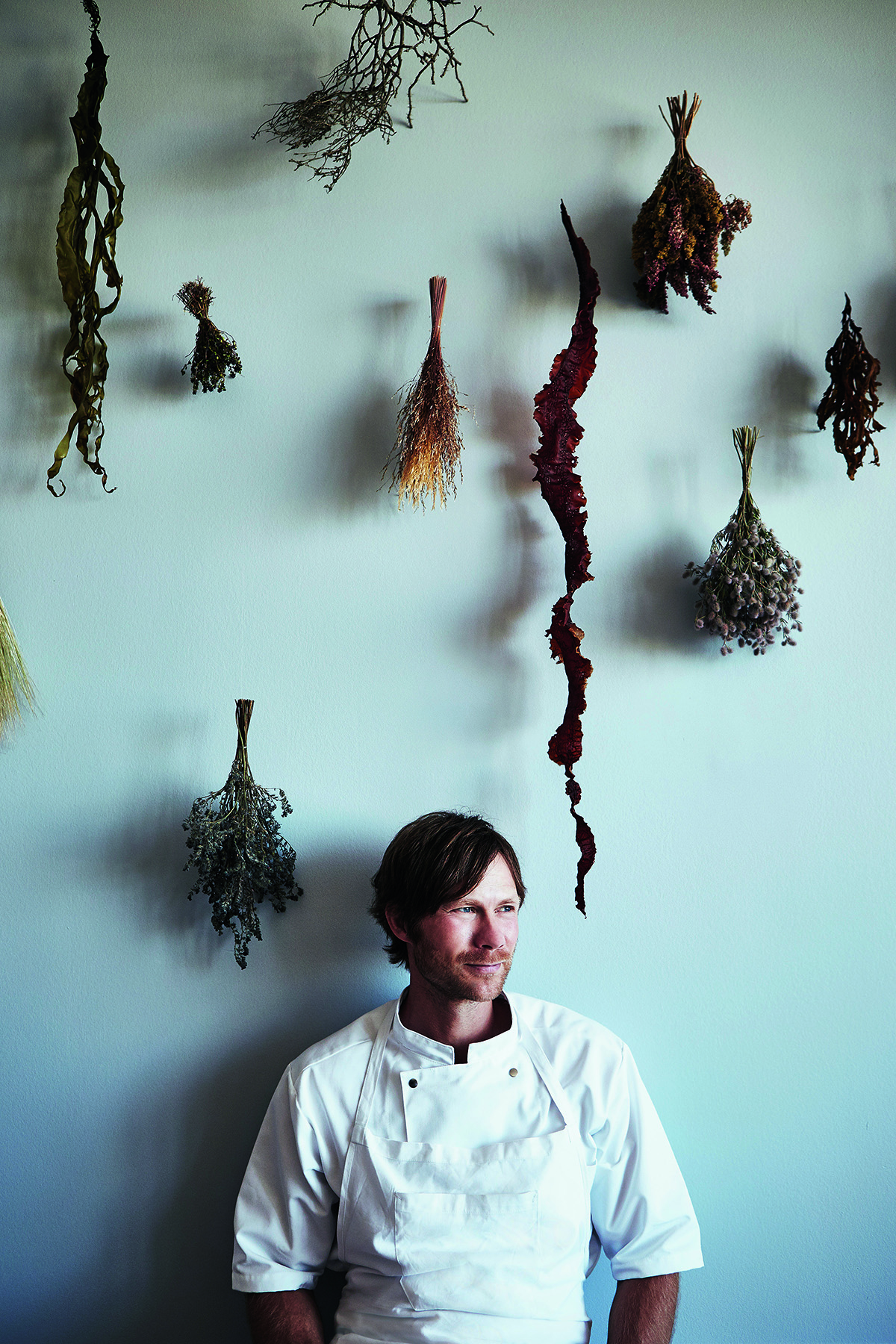 A chef in an apron standing by a wall with geranium hanging by him