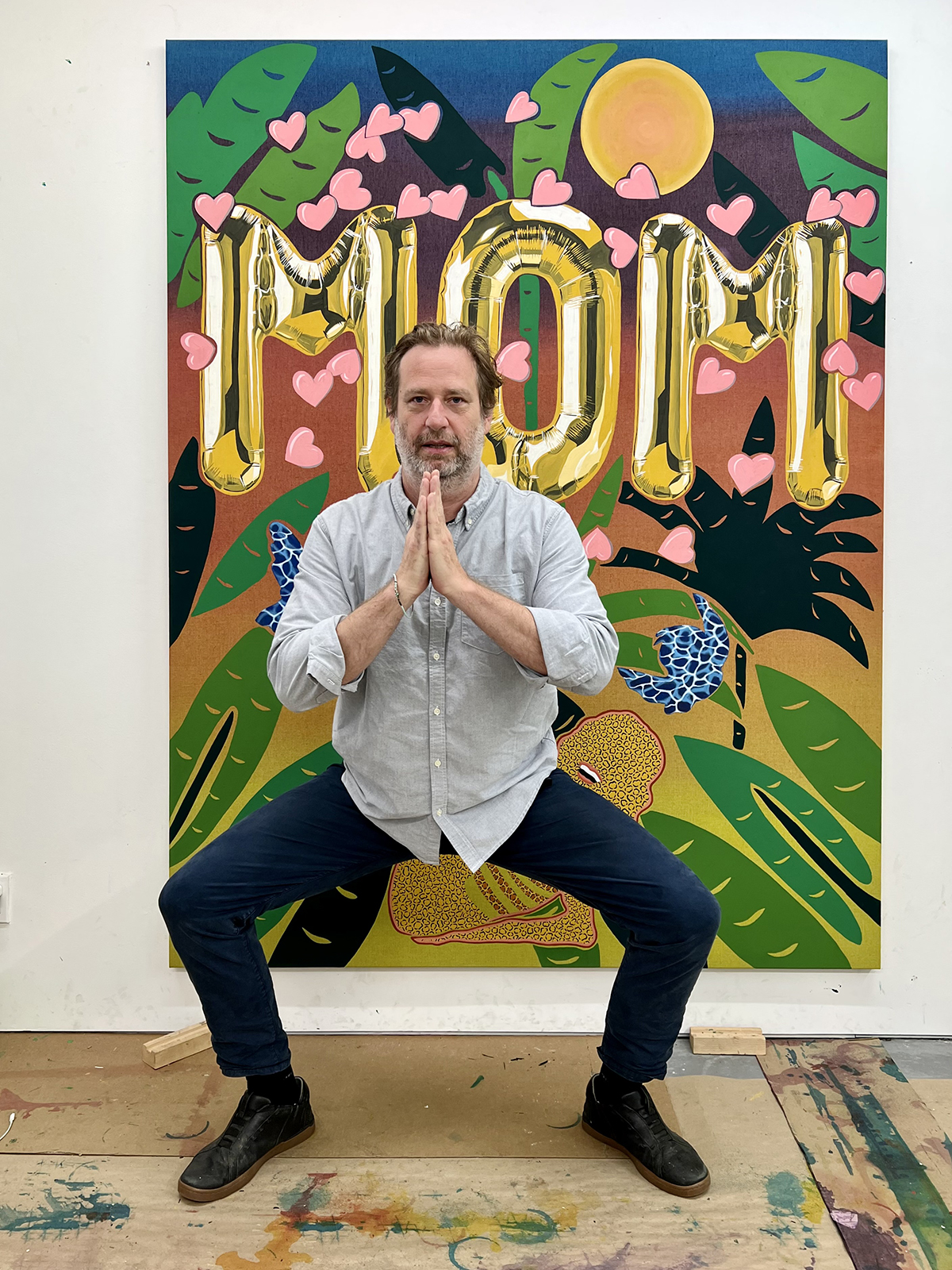 A man squatting in front of a painting that says Mom