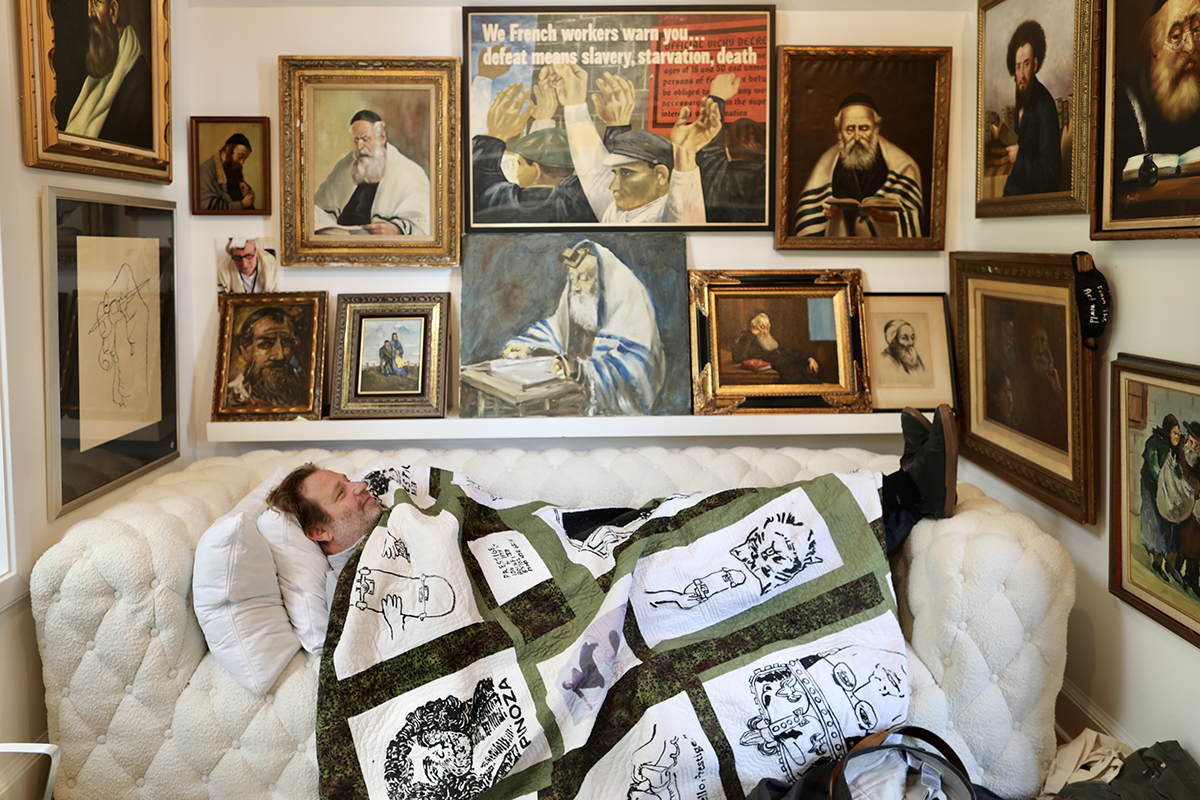 A man lying on a sofa wearing a blanket with peoples faces on it surrounded by pictures of rabbis