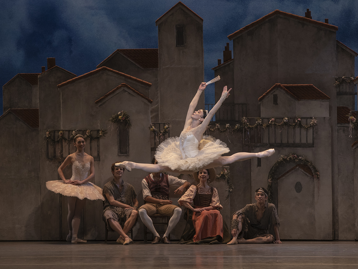 A ballerina wearing a tutu doing a jump in the air and people on the stage watching