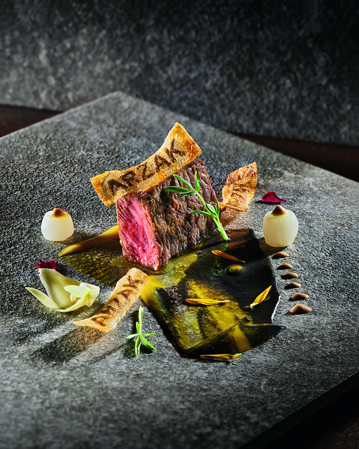 A steak on a plate with a cracker that has the words ARZAK burnt into it