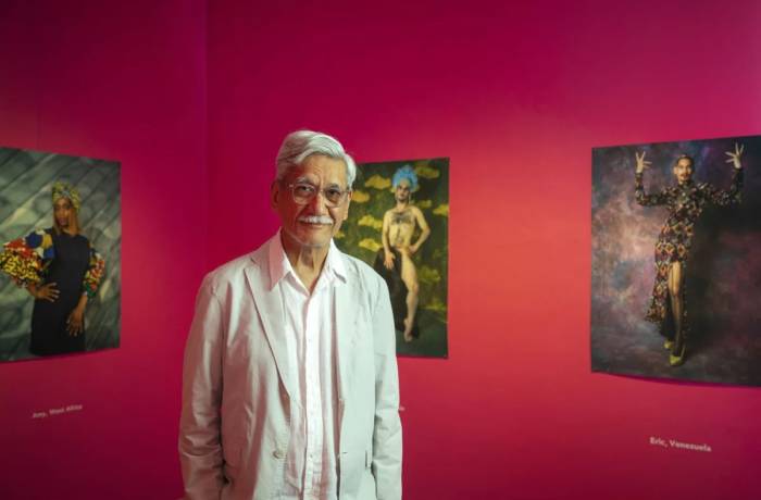 An old man standing in front of a pink wall with framed photographs on the walls