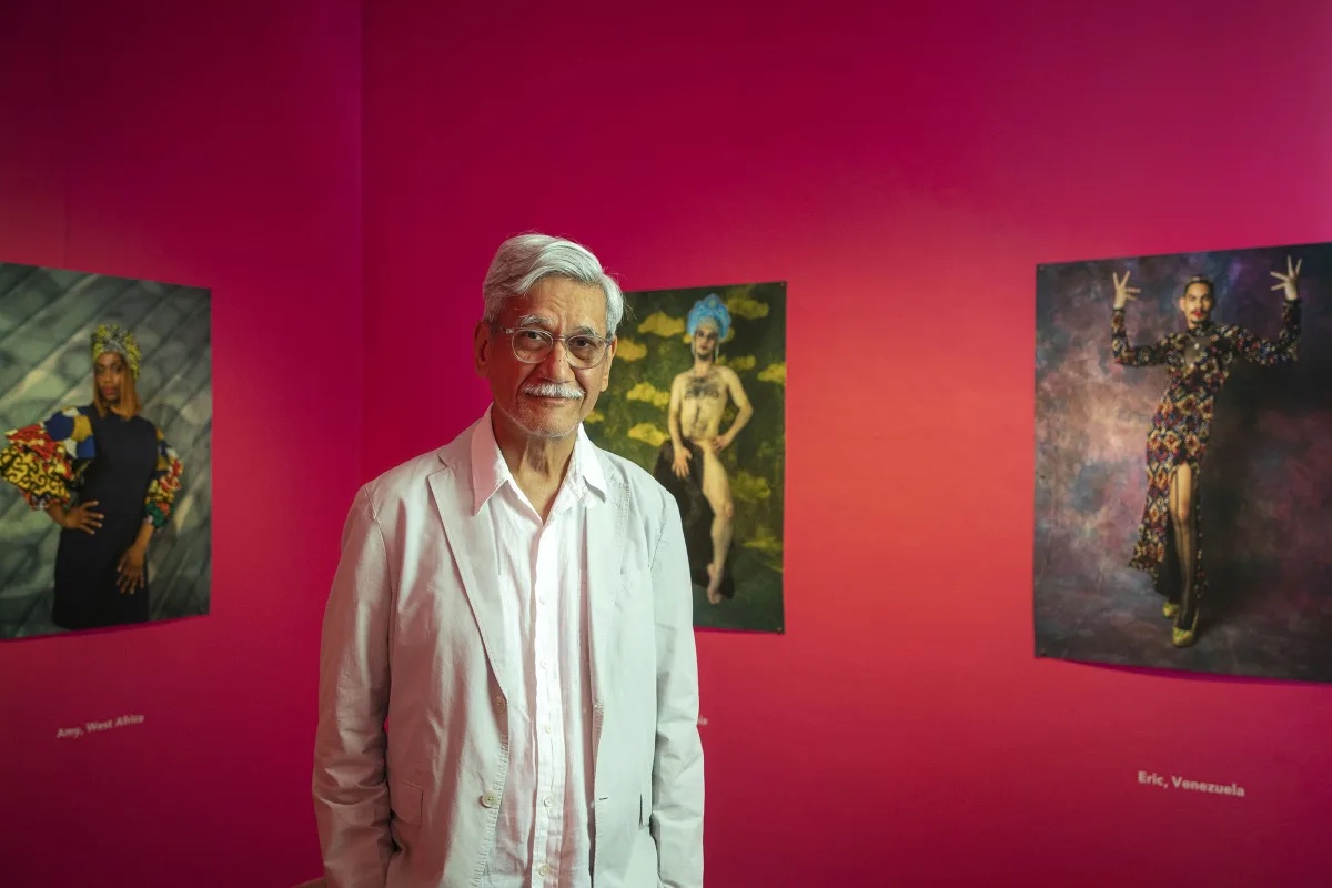 An old man standing in front of a pink wall with framed photographs on the walls