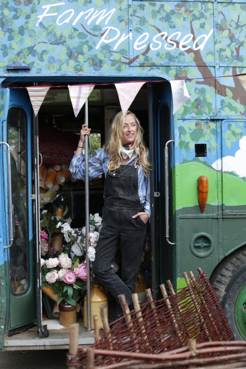 A woman in black dungarees and a blue shirt standing on a greed bus decorated with flags