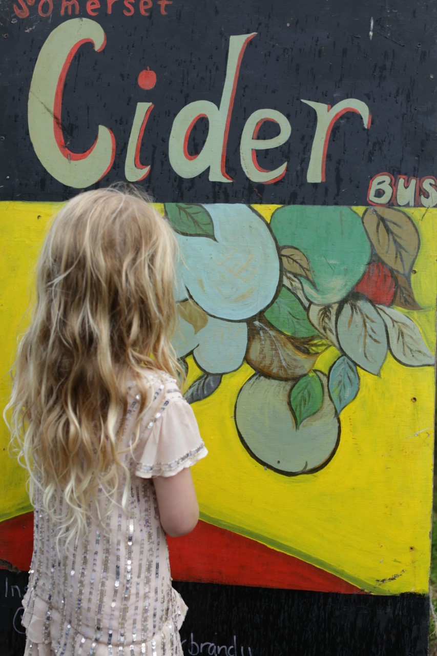 A girl with blonde hair looking at a yellow, green and blue, cider sign