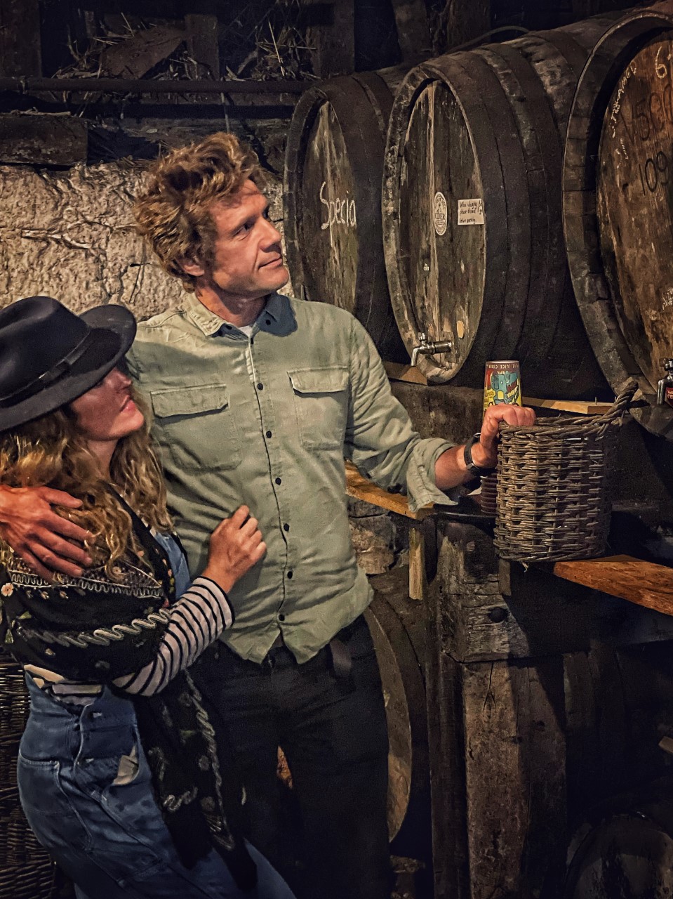 A man in a green shirt with his arm around a woman wearing a hat standing by a distiller in a cellar