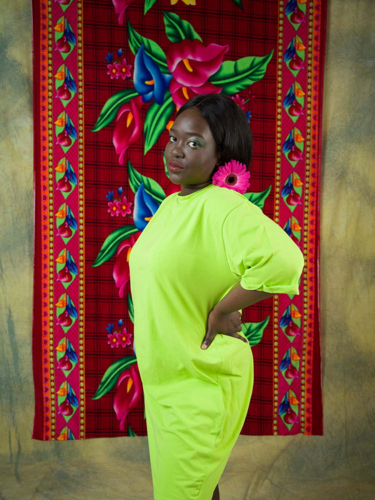 A woman in a neon yellow dress standing in front of a red tapestry with pink and blue flowers on it