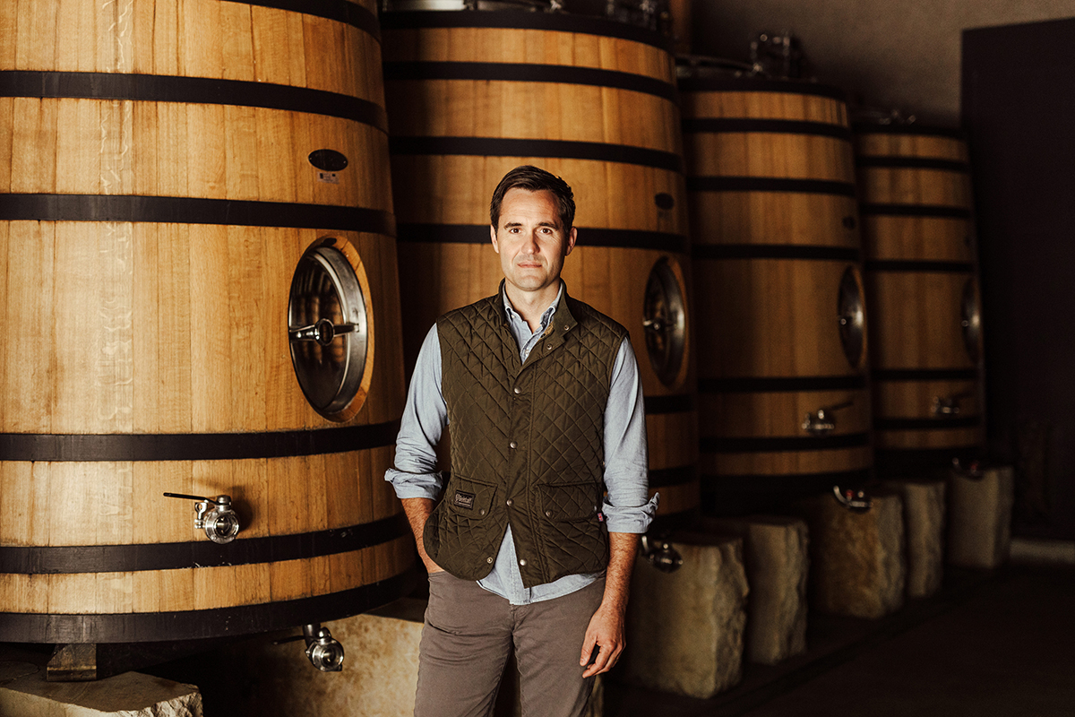 a man in a brown gilet and blue shirt standing in front of barrels of wine