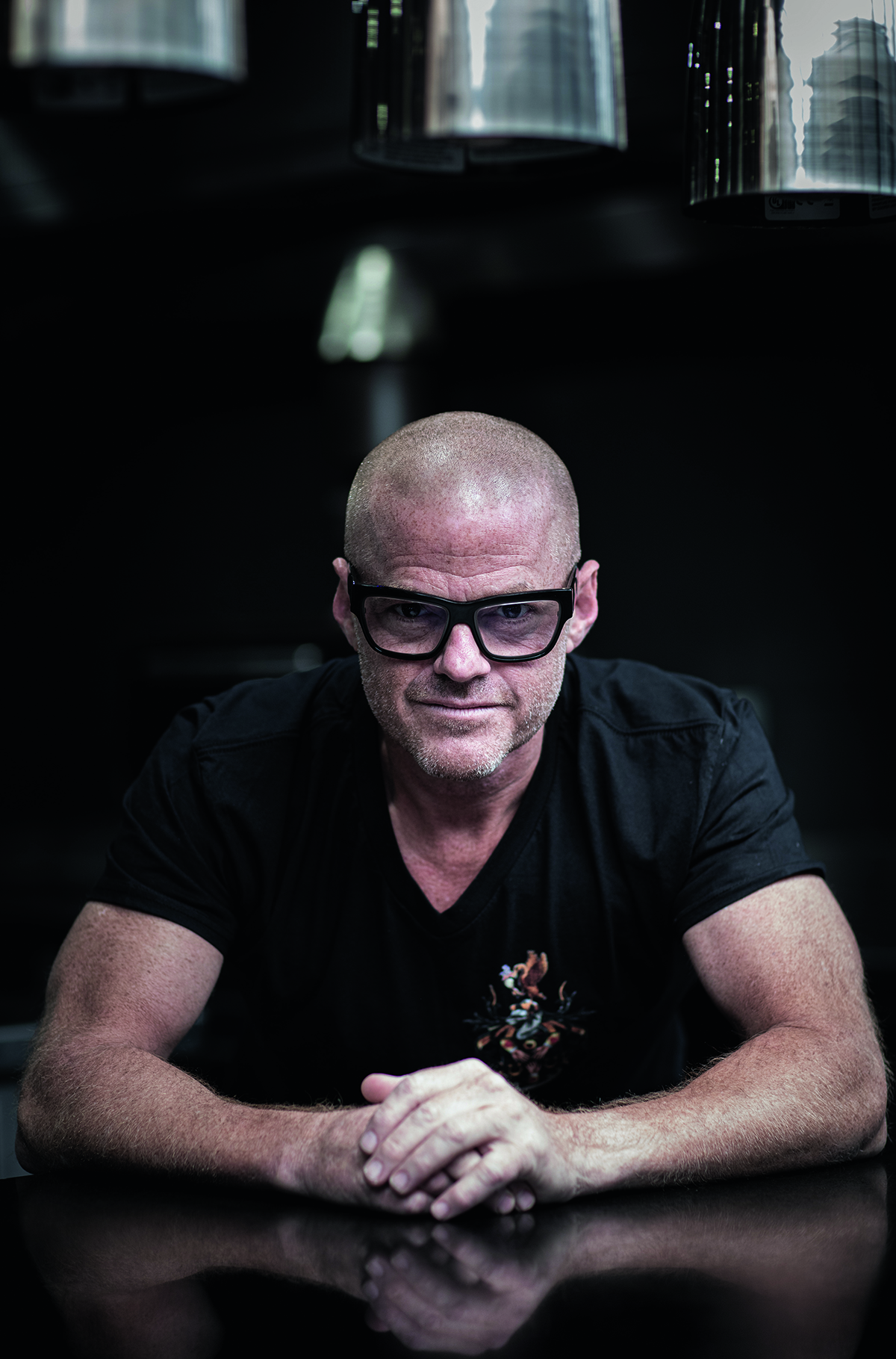 A bald man wearing a v neck black t shirt and glasses staring at the camera