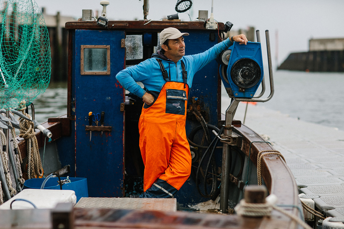 A man standing on a fishing boat wearing an orange jumpsuit and blue top