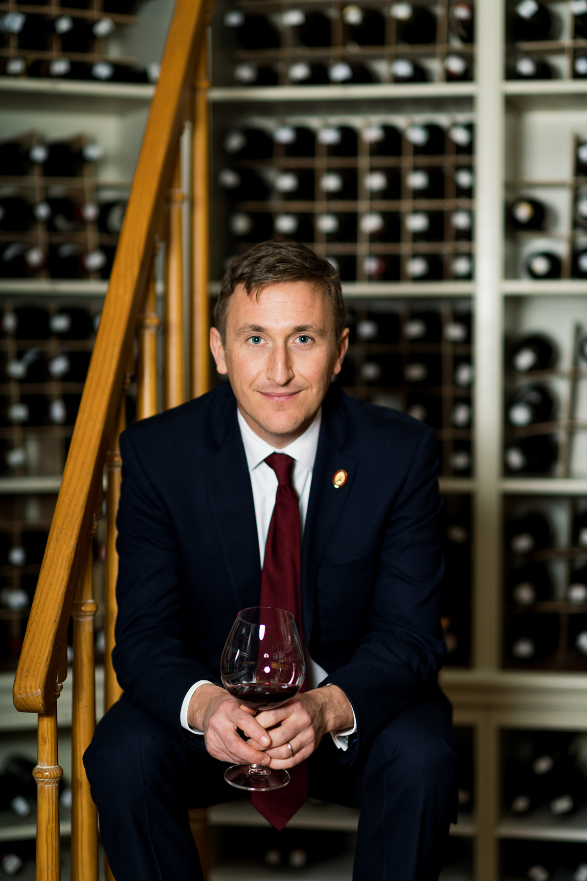 A man in a suit and red wine sitting with a glass of red wine in front of bottles of wine