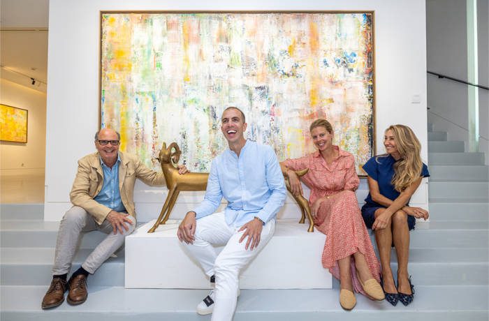 a man in a blue shirt and white jeans sitting next two women in a blue and a red dress sitting in front of a painting