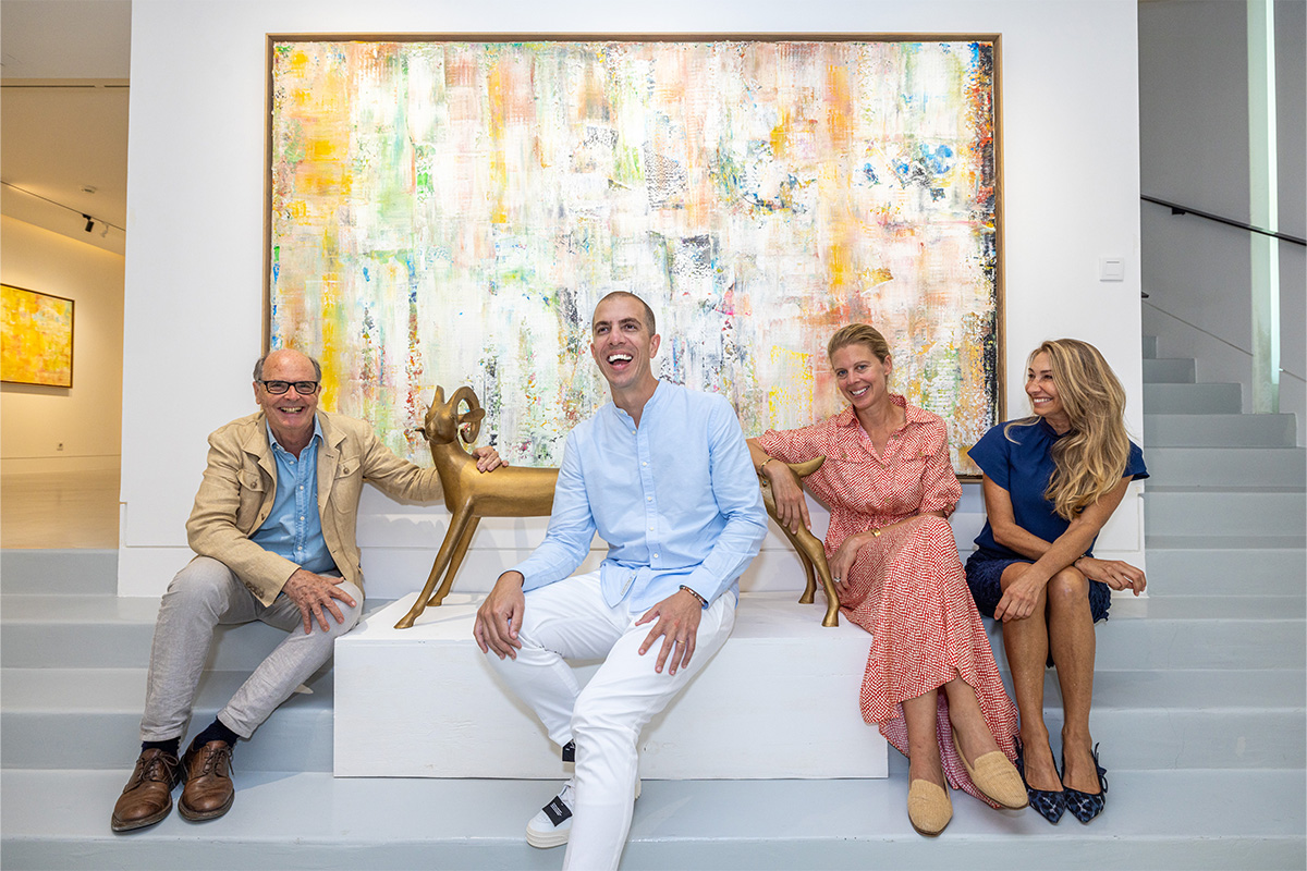 a man in a blue shirt and white jeans sitting next two women in a blue and a red dress sitting in front of a painting