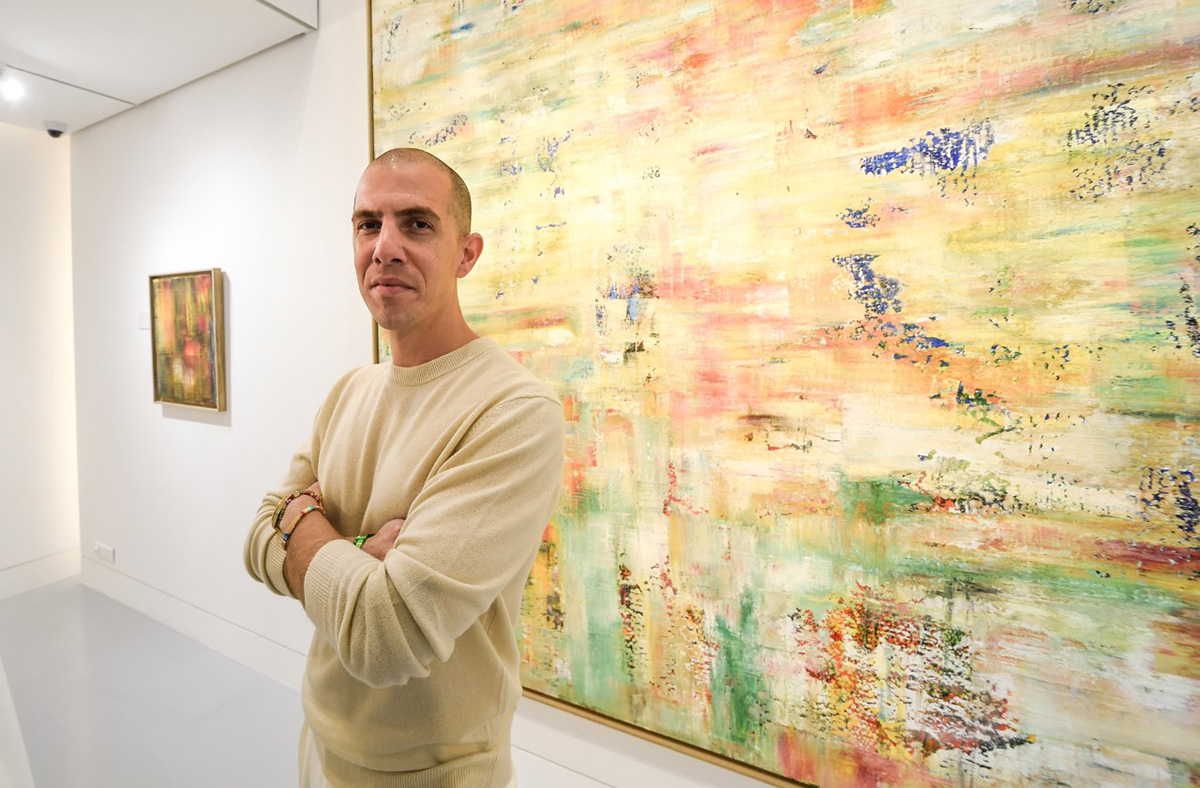 A man in a cream sweater with his arms crossed standing by a yellow painting