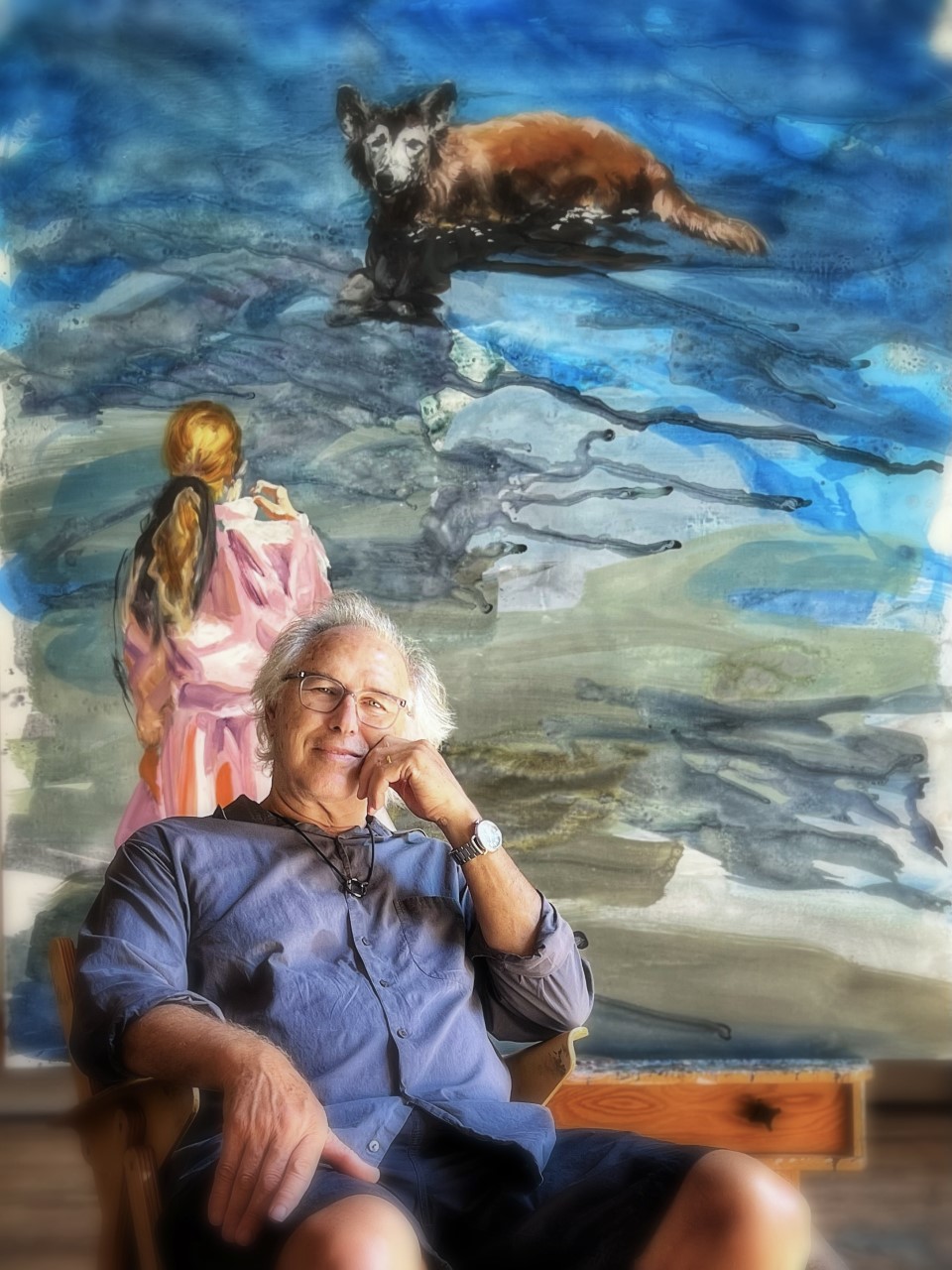 a man in a blue shirt sitting in front of a painting of a girl in a pink dress looking at a dog