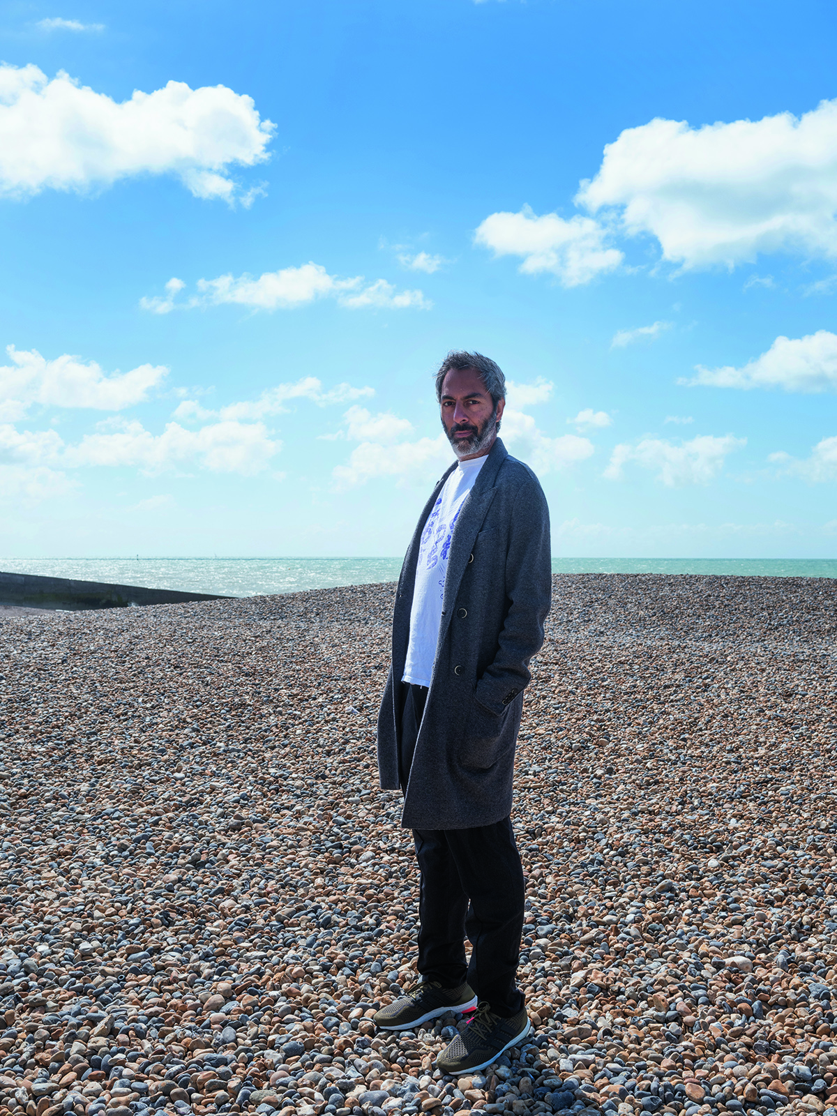 A man standing on a pebbled beach wearing a white t-shirt, black jeans and a long coat 