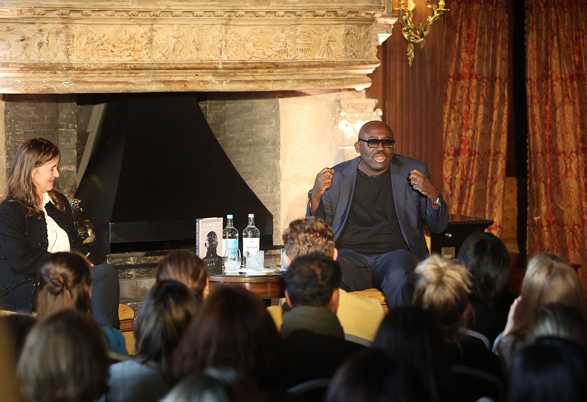 Edward Enniful wearing glasses, a grey blazer, black t-shirt and jeans giving a talk to an audience sitting on a chair