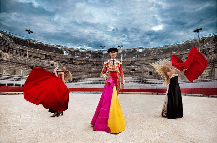 A man in a bullring holding a pink and yellow flag with women on either side of him holding red flags