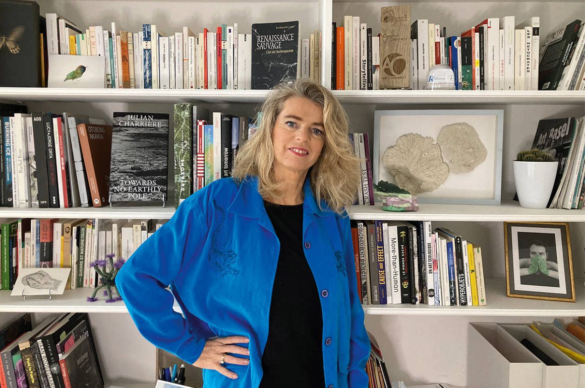 A blonde woman in a black top and blue shirt standing by a book shelf with her hand on her hip