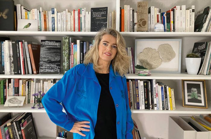 A blonde woman in a black top and blue shirt standing by a book shelf with her hand on her hip