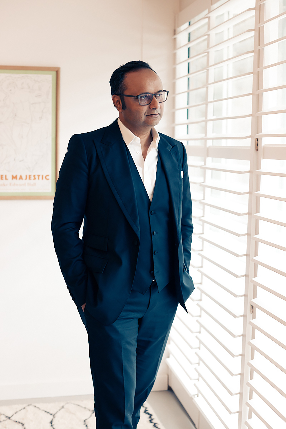 A man wearing a navy suit and white shirt standing by a window 