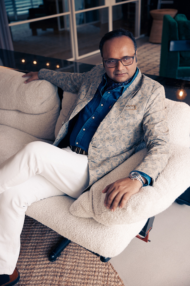 a man sitting on a cream sofa wearing a cream jacket, white trousers and a denim shirt