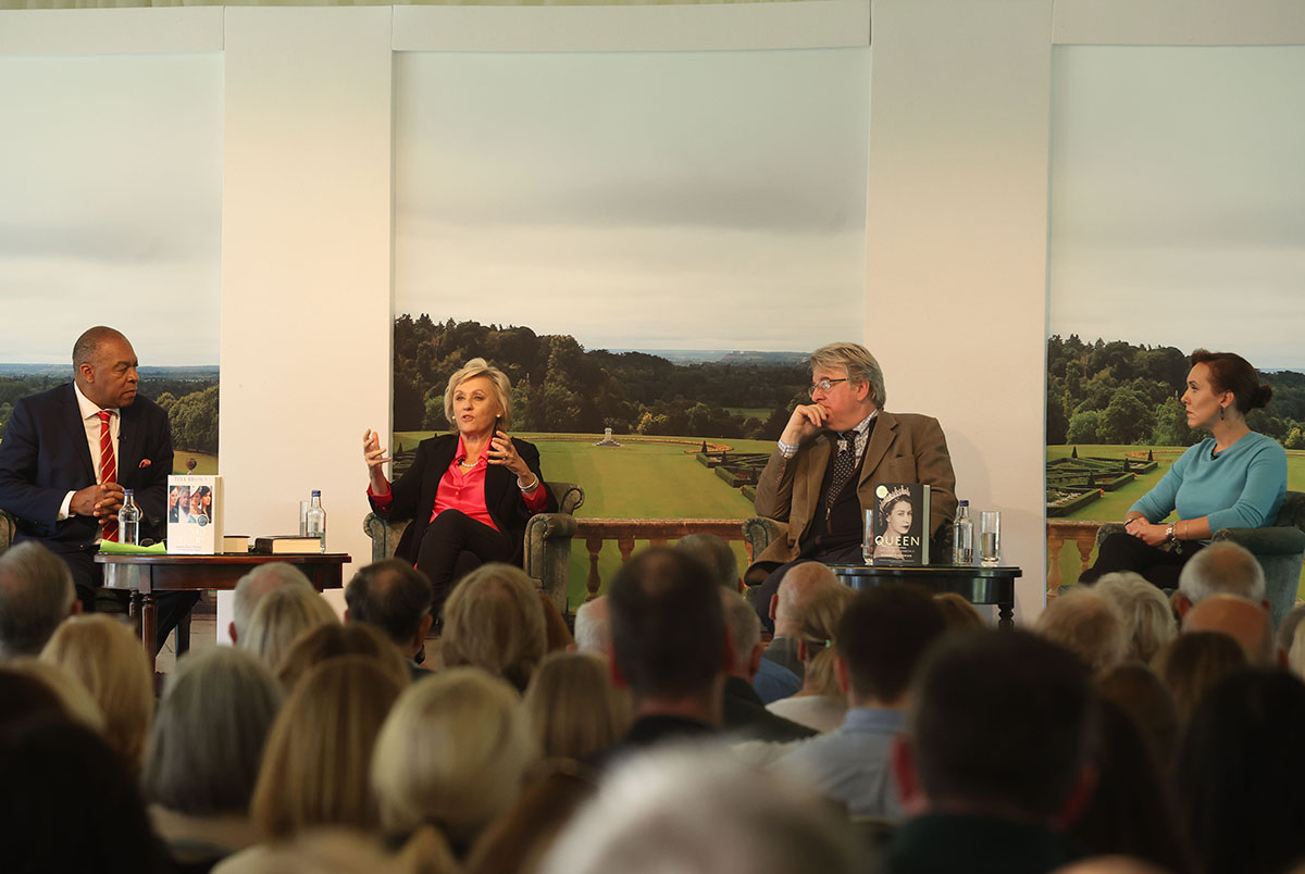 A woman in a pink shirt and black suit hosting a talk with a man wearing a brown suit