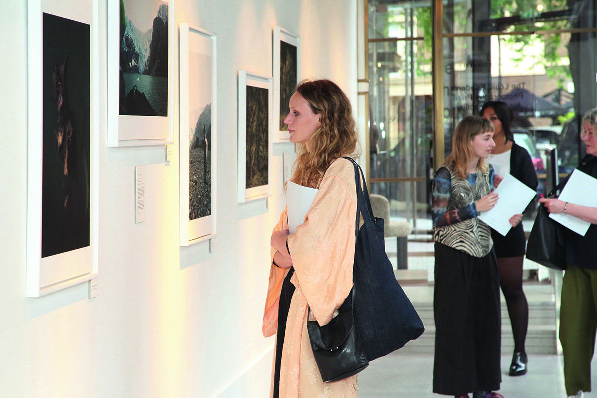 A woman wearing a peach coloured coat and black bag looking at a picture on a wall