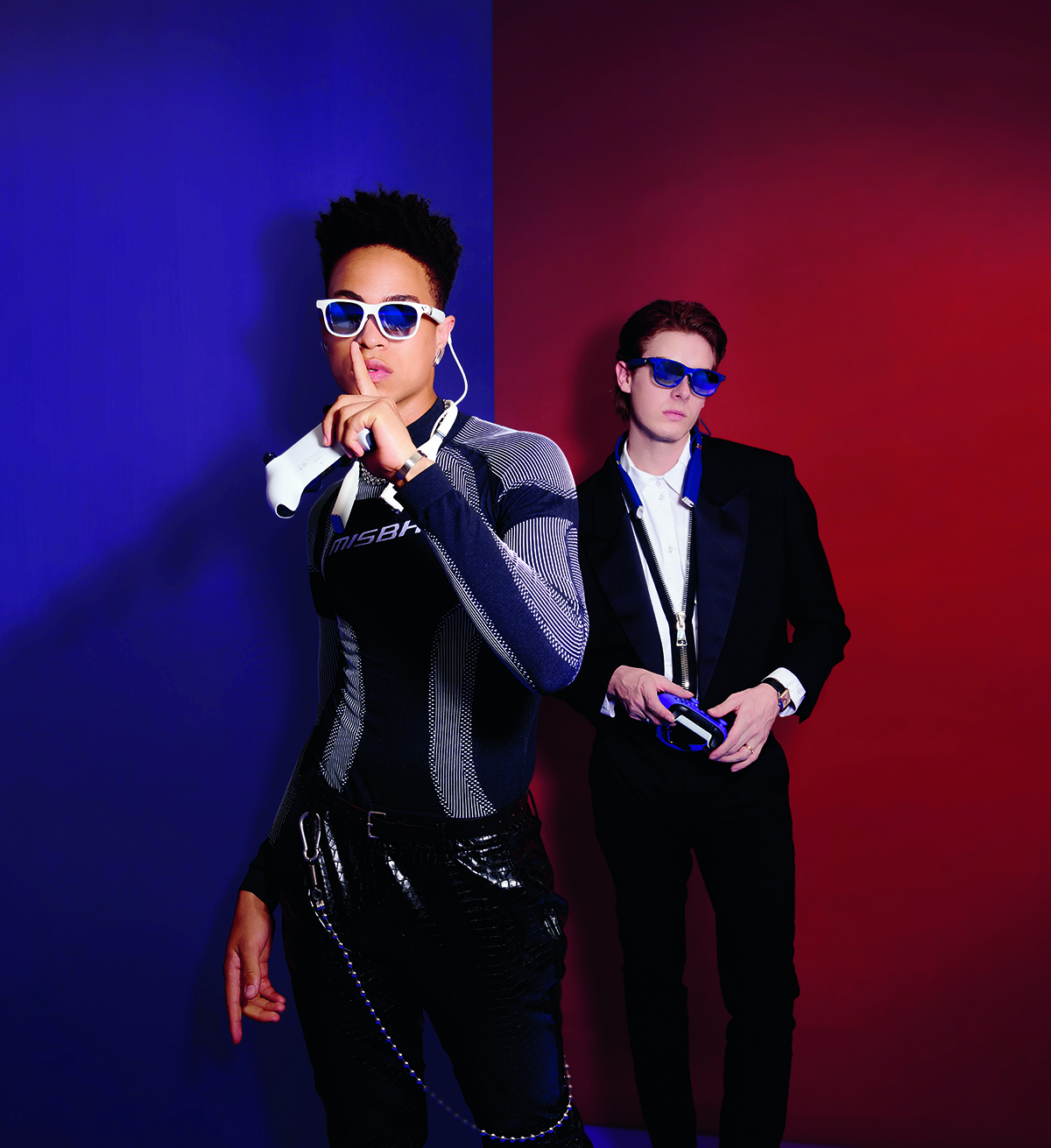 purple and red background with a model with his finger to his lips in a leather outfit wearing glasses and a man wearing a suit in the background also wearing glasses
