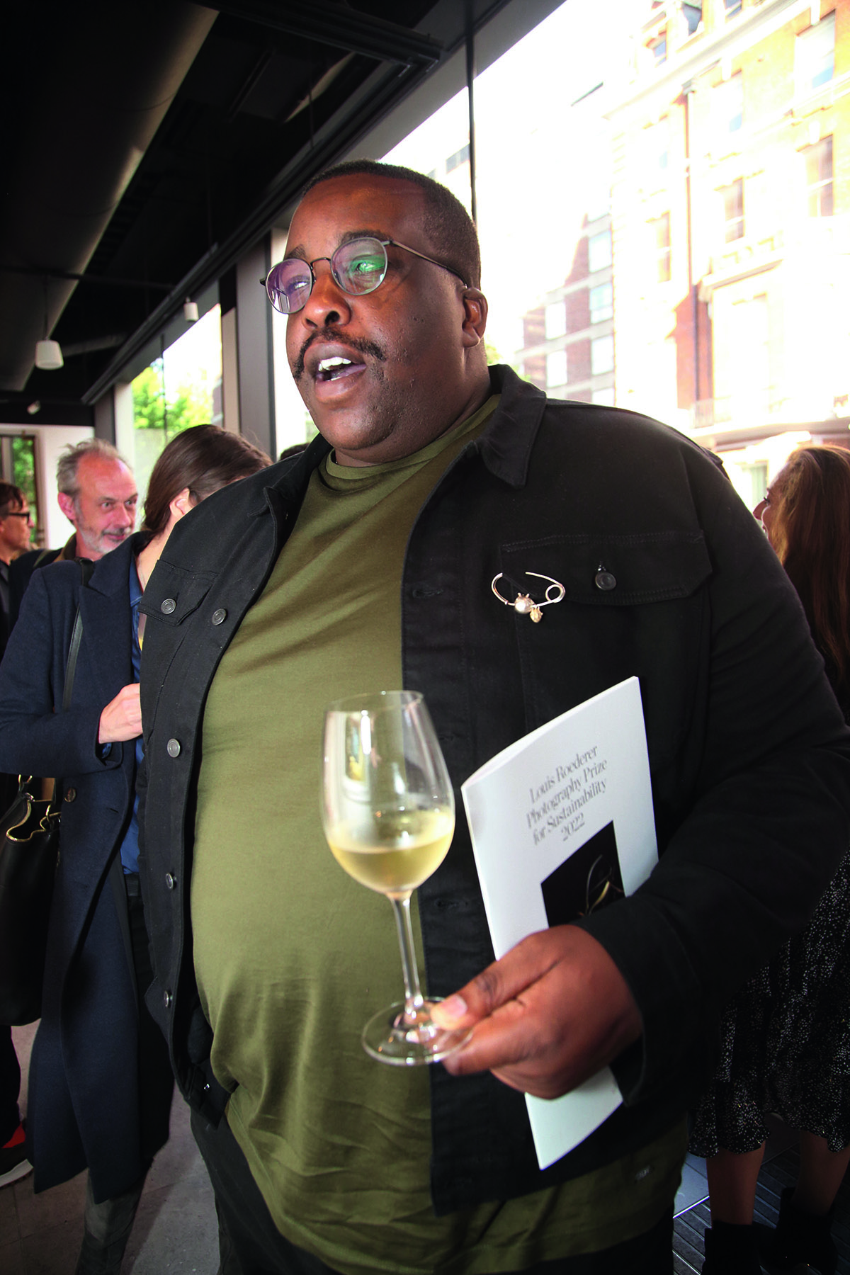 A man holding a champagne glass wearing a green t shirt and black jacket