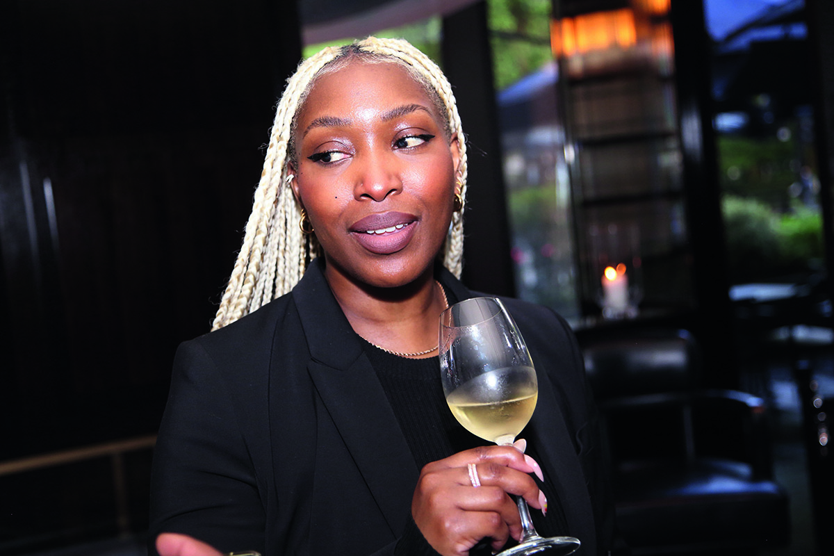 A woman with blonde cornrows wearing black holding a champagne glass