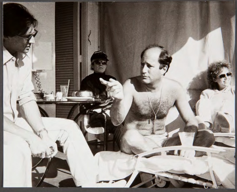 black and white photo of a topless man sitting with another man at a table 