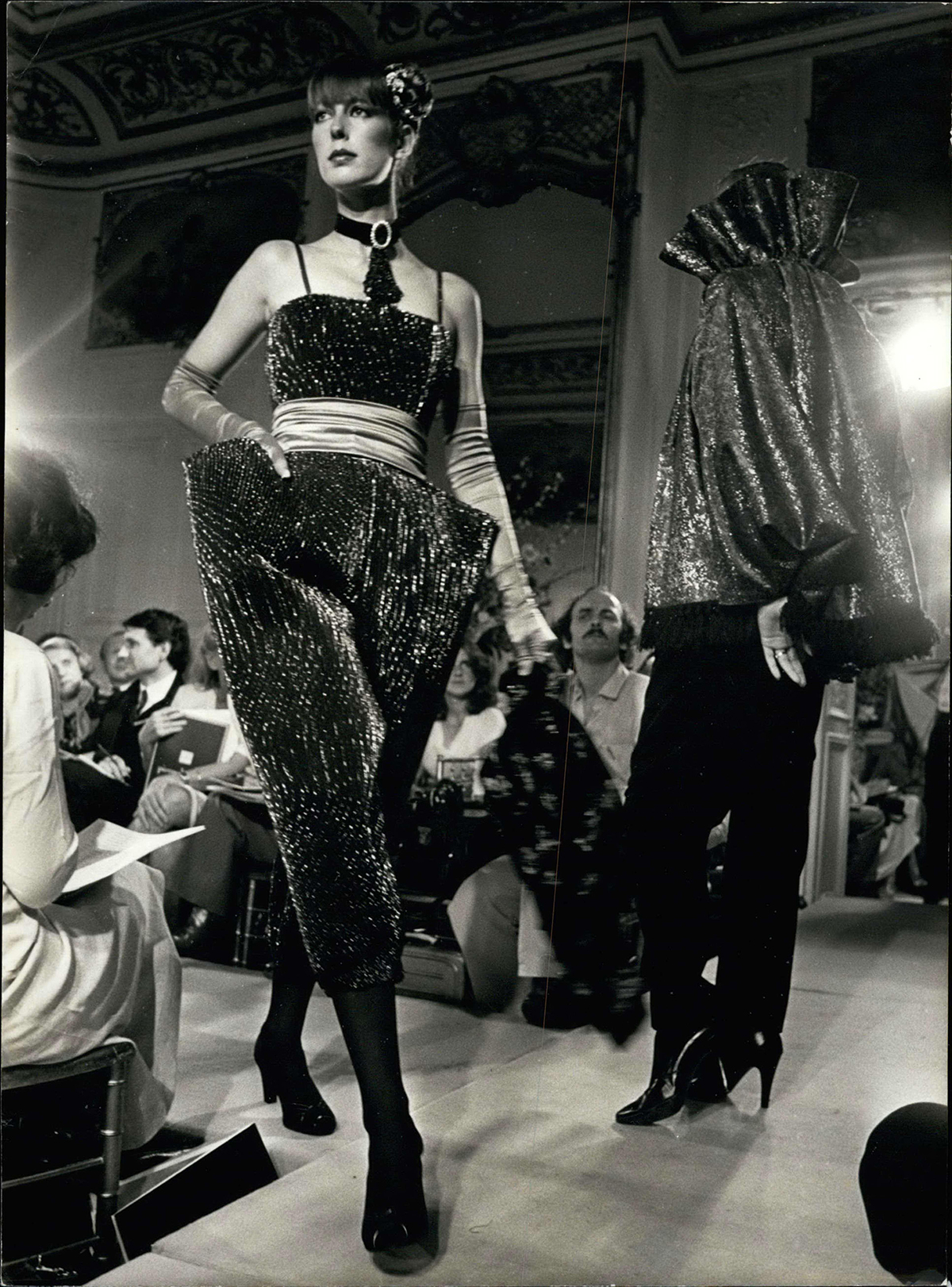 A black and white photo of a model on a catwalk wearing a black vest and large angled trousers