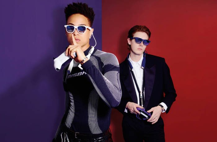 purple and red background with a model with his finger to his lips in a leather outfit wearing glasses and a man wearing a suit in the background also wearing glasses