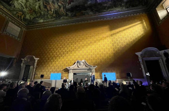 A talk being hosted with an audience in a room with a marble fireplace and yellow wallpaper and a painted ceiling