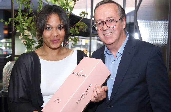 Woman in a white and black top holding a pink case of champagne standing next to a man in a blue shirt and black blazer