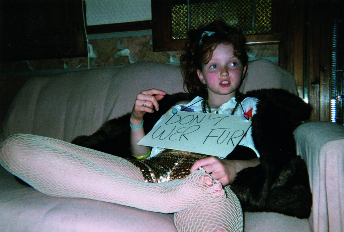 A child sitting on a sofa with tights on and a sign over her neck that says 'Don't Wer Fur'