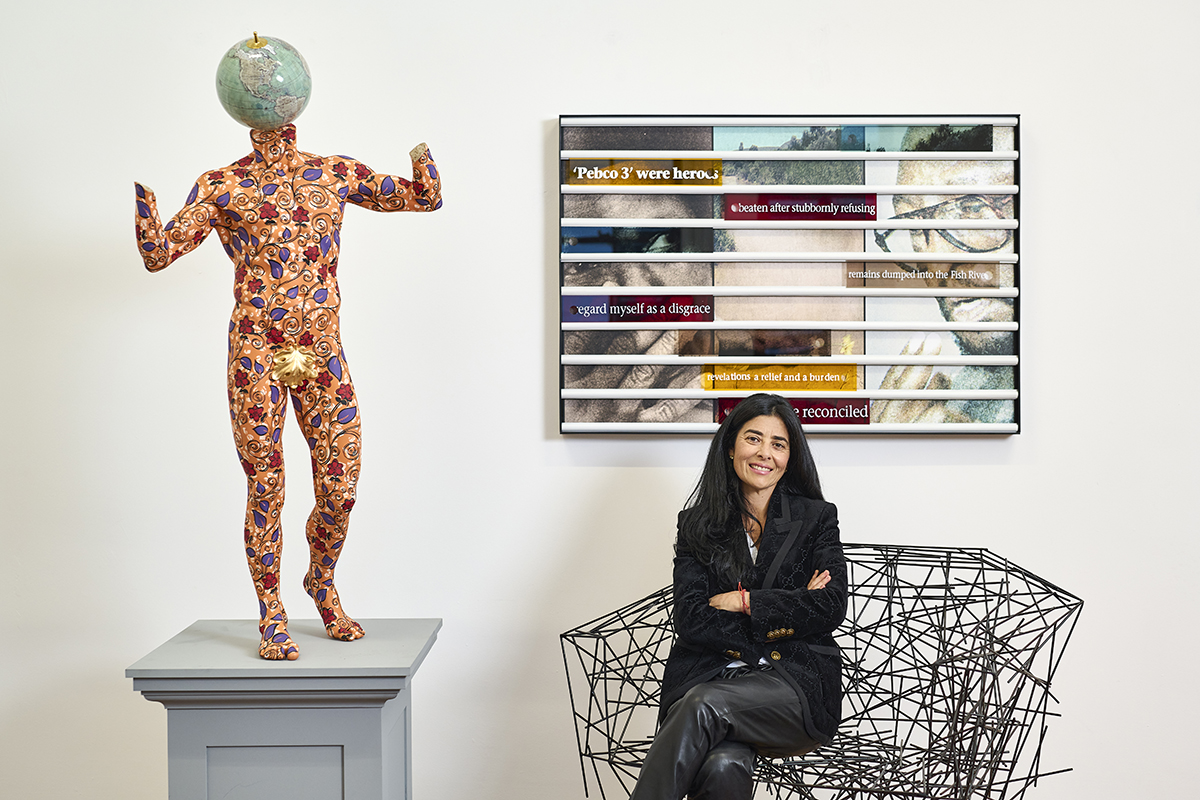 A woman wearing black sitting on a silver chair and a sculpture of a person with a green head and colourful body next to her