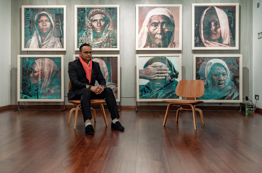 A man wearing a black suit and pink shirt sitting on a chair in front of green portraits hing up on a wall