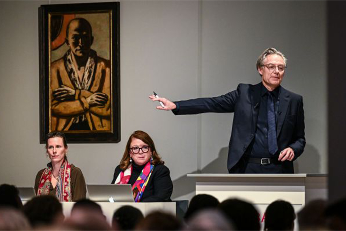 A man showing a painting at an auction with two women beside him