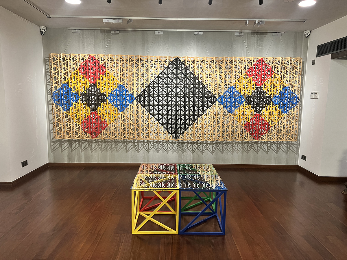 A yellow, blue, red and black wooden clock with cut out shapes hanging on a wall and open sided cubes in blue, yellow, greed and red on the wooden floor