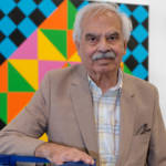 A man wearing a beige jacket and striped shirt standing in front of a geometric painting