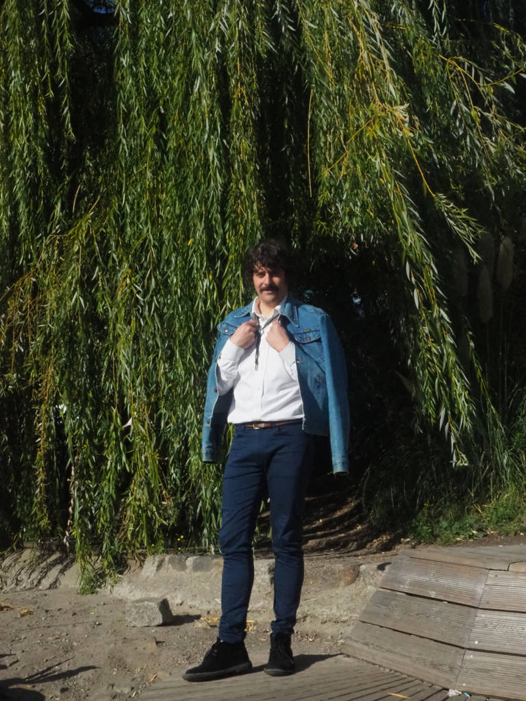 A man wearing a white shirt, jeans and a denim jacket over his shoulders standing by a willow tree 
