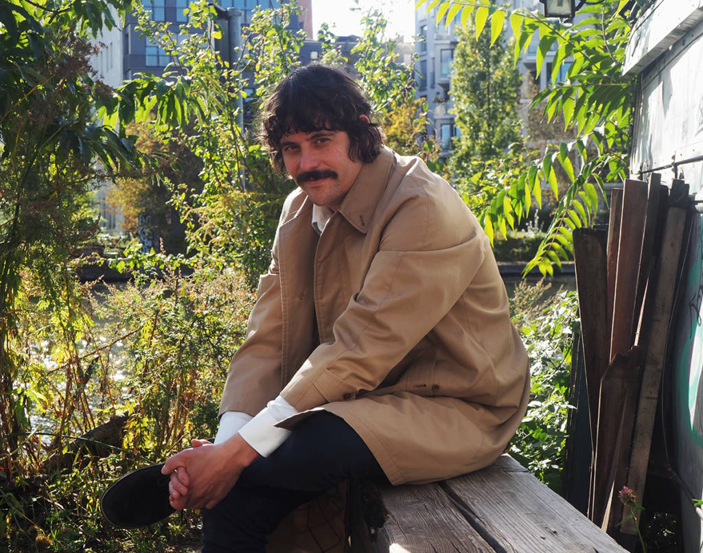 A man sitting on a bench next to plants wearing a trench coat